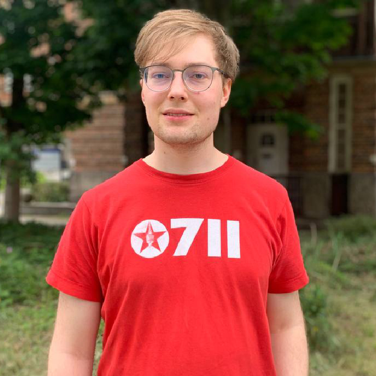 Bild zeigt einen jungen Mann vor einem Backsteinhaus und einer Wiese. Er wurde im Potraitmodus fotografiert und trägt ein rotes T-Shirt